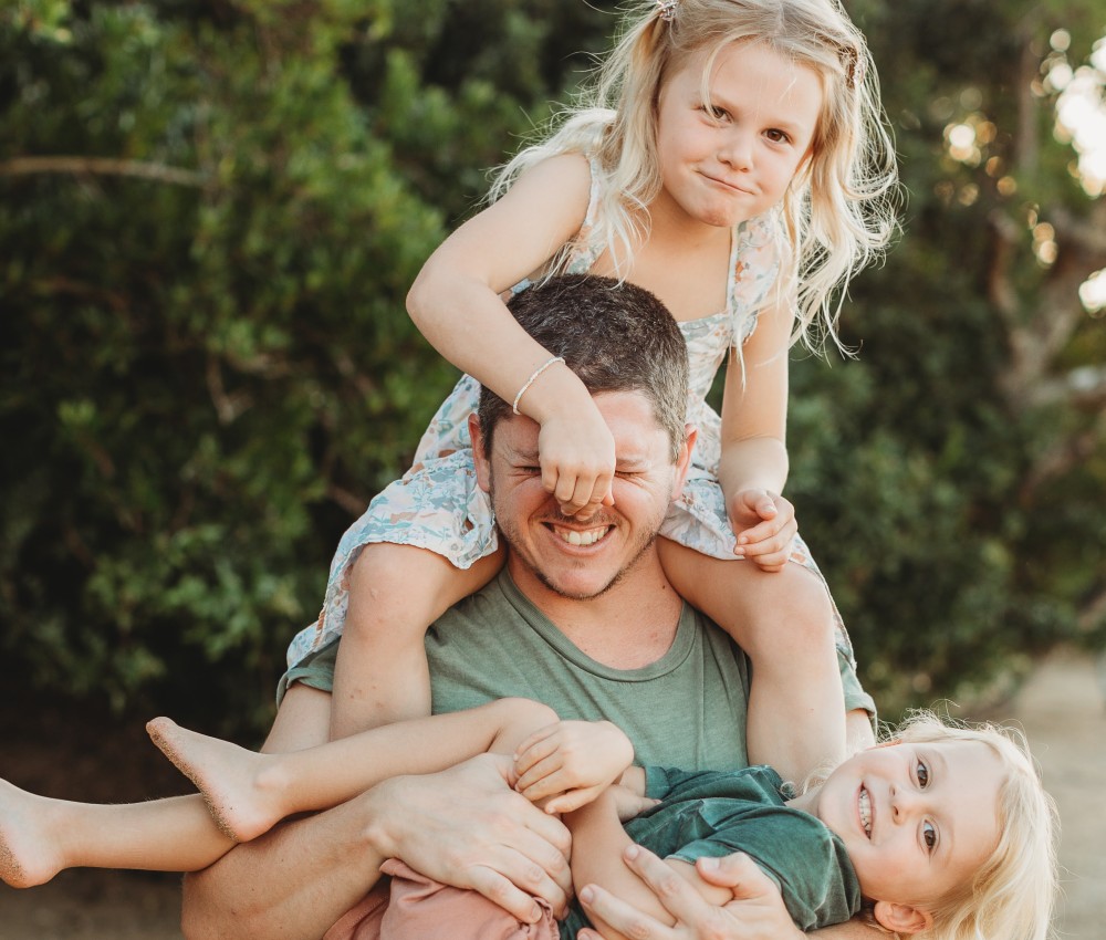 Brad laughing outdoors with his children climbing over him.